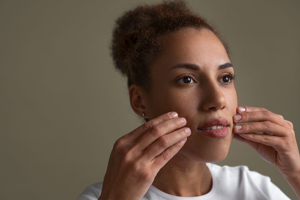 A imagem mostra uma mulher com cabelo crespo, usando uma camiseta branca, realizando um exercício de yoga facial. Ela está com as mãos posicionadas nas bochechas, suavemente apertando a pele, em um gesto que parece ser parte de uma massagem facial. A expressão da mulher é concentrada, mas relaxada, sugerindo que ela está focada no exercício. O fundo é neutro, com uma tonalidade suave, que não distrai a atenção do rosto da mulher e da técnica que ela está aplicando para manter a pele jovem e saudável.