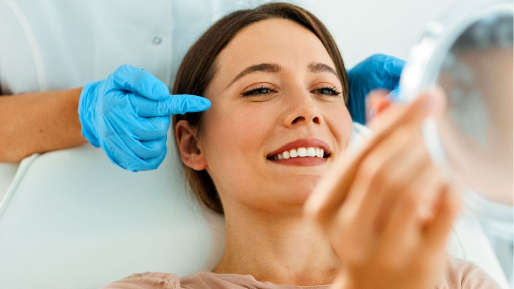 A imagem mostra uma mulher sorrindo enquanto segura um espelho em uma das mãos. Ela está em um ambiente clínico, com um profissional ao fundo, usando luvas azuis, apontando para sua testa, provavelmente preparando-se para aplicar um tratamento estético. A mulher parece relaxada e satisfeita com o procedimento, enquanto observa os resultados no espelho. O ambiente transmite um ar de cuidado e atenção, evidenciando um procedimento estético não invasivo e focado no bem-estar da paciente.