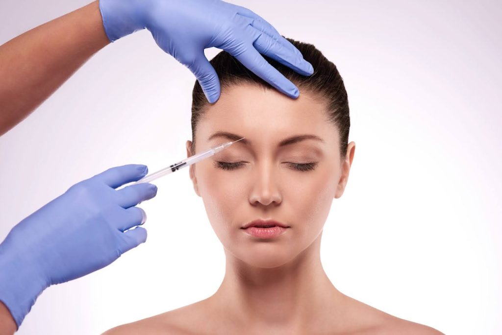 A imagem mostra uma mulher recebendo a aplicação de toxina botulínica (Botox) na testa. Ela está com os olhos fechados, aparentando estar relaxada enquanto um profissional, usando luvas azuis, aplica a substância com uma seringa na região entre as sobrancelhas. O ambiente é clínico e profissional, com um fundo neutro que destaca o procedimento estético sendo realizado. A imagem transmite um senso de cuidado e precisão no tratamento, com foco no rejuvenescimento facial.