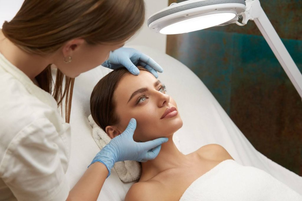 A imagem mostra uma mulher recebendo um tratamento estético em um ambiente clínico. Ela está deitada, com um lençol cobrindo o corpo, e parece relaxada enquanto uma profissional, usando luvas azuis, está aplicando cuidados em seu rosto, provavelmente preparando a área para algum procedimento. A luz suave de um equipamento de tratamento ilumina o local, e o foco está no cuidado detalhado da pele ao redor da área dos olhos e testa. O ambiente transmite um ar de profissionalismo e atenção ao bem-estar do paciente.