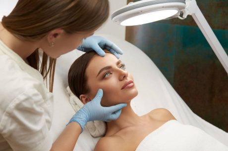 A imagem mostra uma mulher recebendo um tratamento estético em um ambiente clínico. Ela está deitada, com um lençol cobrindo o corpo, e parece relaxada enquanto uma profissional, usando luvas azuis, está aplicando cuidados em seu rosto, provavelmente preparando a área para algum procedimento. A luz suave de um equipamento de tratamento ilumina o local, e o foco está no cuidado detalhado da pele ao redor da área dos olhos e testa. O ambiente transmite um ar de profissionalismo e atenção ao bem-estar do paciente.